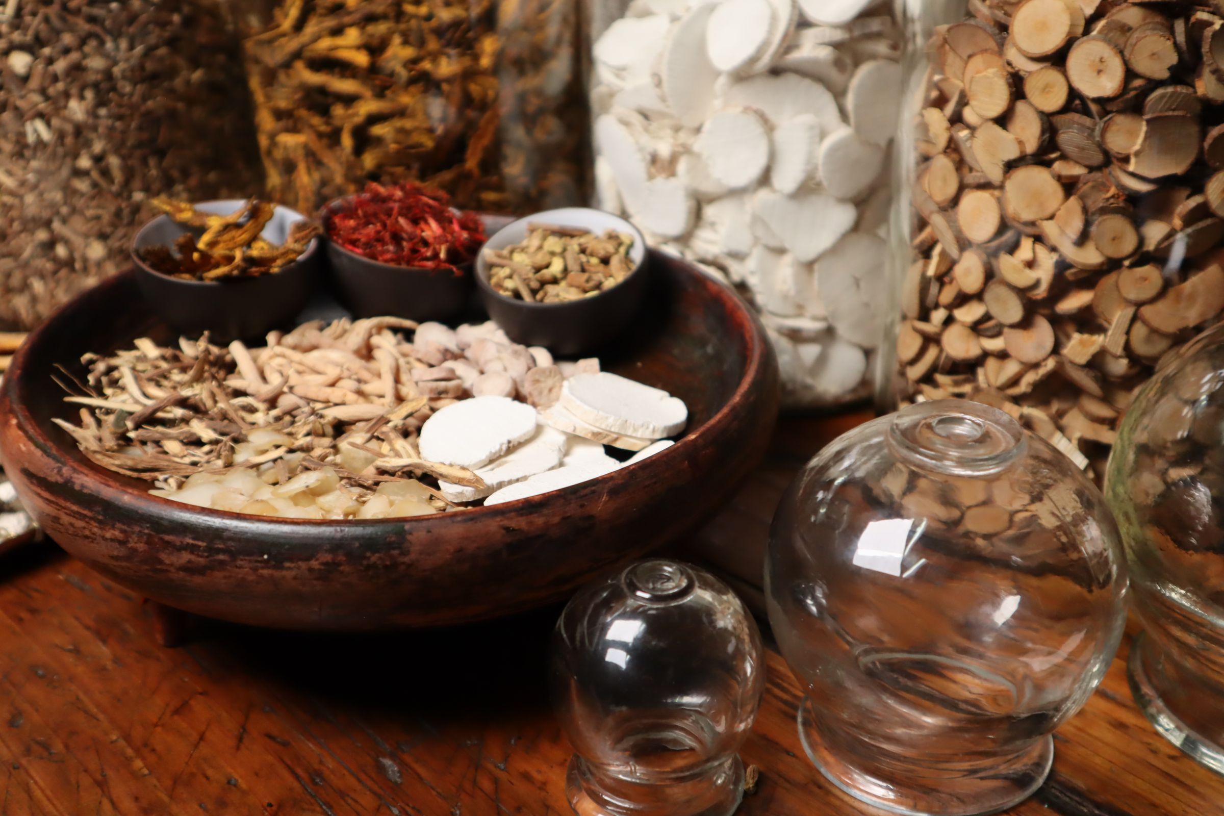 Dried roots, seeds and herbs in a wooden bowl beside glass jars